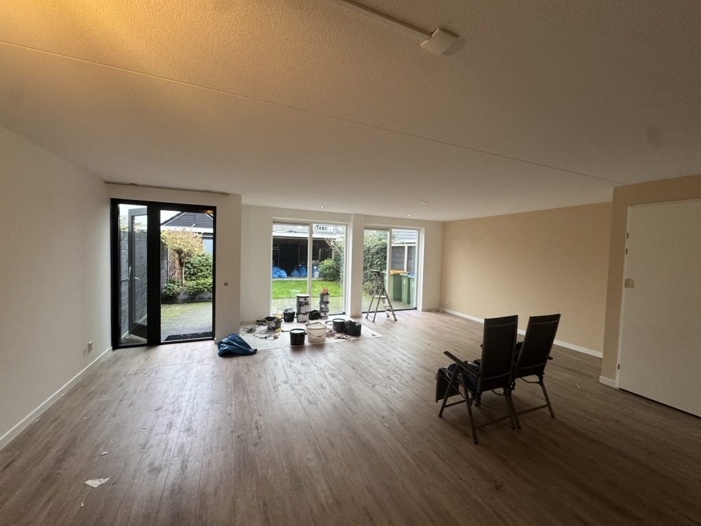 Lege doorzonwoonkamer van een rijtjeshuis met houten vloer, lichte wanden en verfemmers bij de tuindeuren